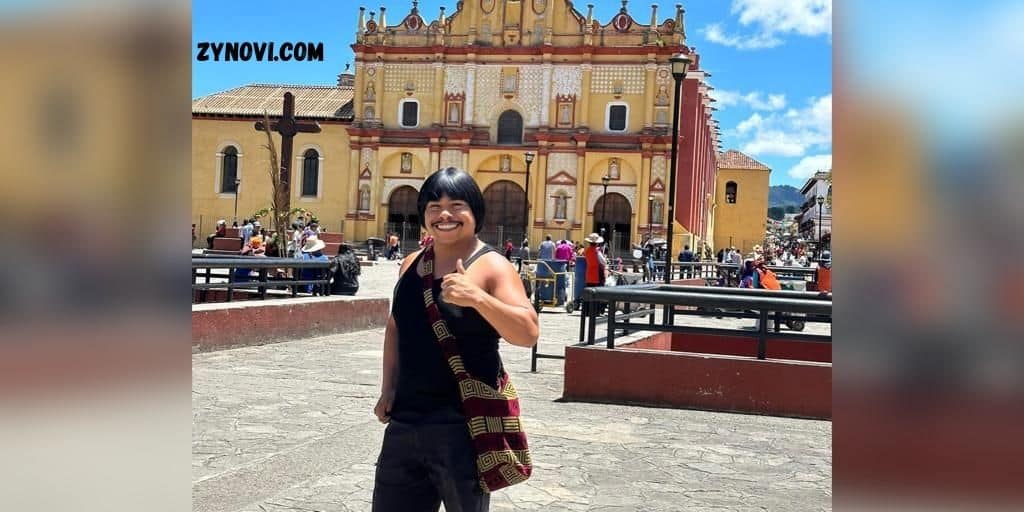 Chaparro Chuacheneger poses in a black shirt and hat before a church, showcasing a blend of style and reverence.